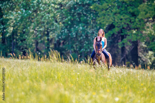 Young woman with brunette long hair rides bareback with her brown horse across a summer meadow, dressed in a blue tank top and riding pants with boots.