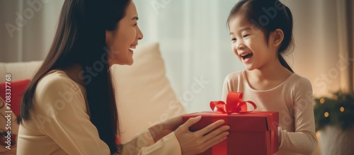 Asian girl surprising happy mother with gift on couch at home.
