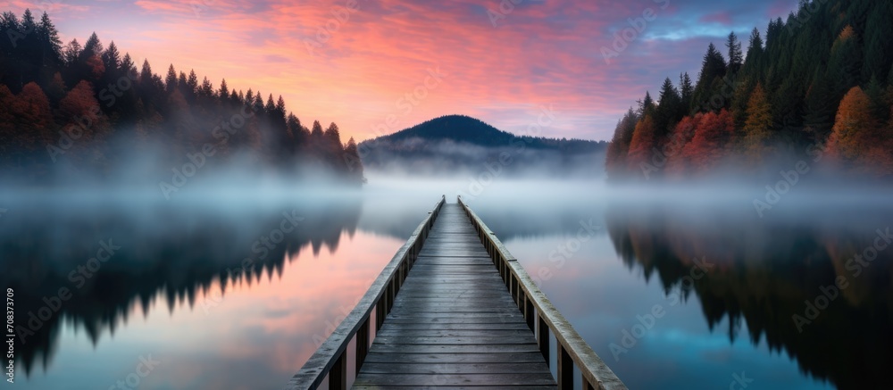 A wooden pier extends into a foggy lake, reflecting trees, tranquil water, and a colorful sky.