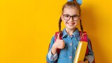 Smiling schoolgirl, 8 years old, showing thumbs up, holding book in her hand with school backpack, yellow background.