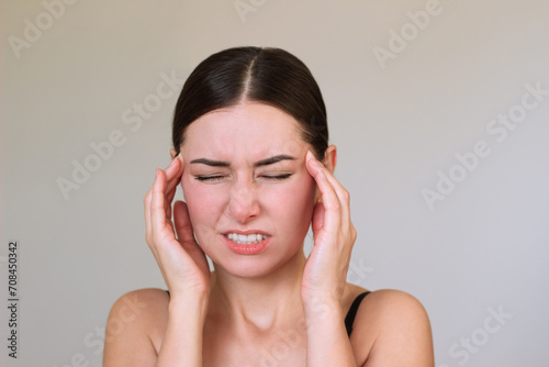 A young Caucasian brunette woman is suffering from severe pain in her head  holding her temples with fingers and with eyes closed  clenching her teeth.