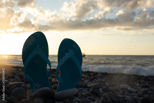 Blue turquoise flip-flops on pebble beach against backdrop of setting sun, sea waves, evening sky. Stones on ocean shore. Concept of holiday, summer vacation, relaxation, resort, relax, rest, tourism