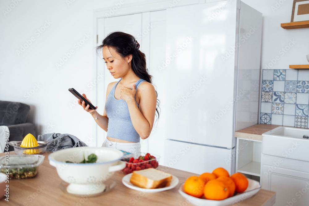 Young attractive Asian woman uses her phone and searches for recipe for dish on Internet. Charming female Korean cooks food in kitchen at home. Young attractive Asian woman uses her phone and searches for recipe for dish on Internet. Charming female Korean cooks food in kitchen at home.