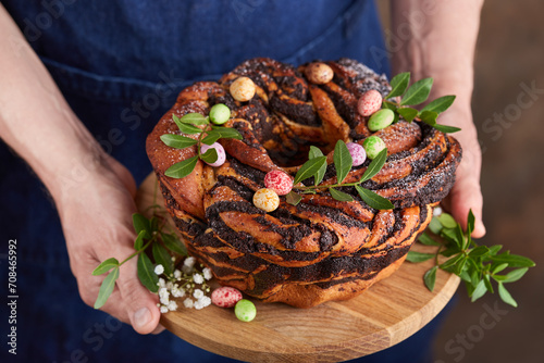 Man in a blue apron holding a freshly baked Easter cake with poppy seed filling and candies in a shape of eggs. Selective focus. Round yeast dough sweet bun. photo