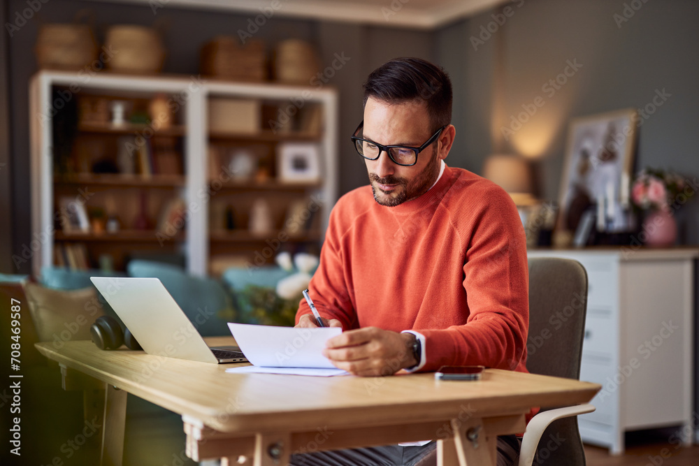 Businessman working from home in an orange sweater. He is signing some documents.