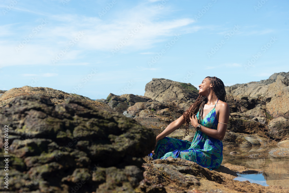 Beautiful woman, in a blue dress, sitting on the rocks with her hand in her braids, sunbathing on the beach looking to the side.