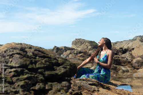 Beautiful woman, in a blue dress, sitting on the rocks with her hand in her braids, sunbathing on the beach looking to the side.