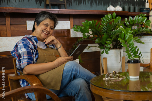 Asian barista female 60s elderly  senior pensioner looking to camera sitting in cozy cafe coffee shop holding modern technology tablet happy with stock market investment  lifestyle retirement women