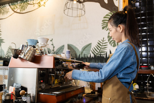 Asian barista female making hot coffee from professional machine standing behind cafe shop counter bar, young waitress worker or small business entrepreneur wearing apron working in eatery cafeteria