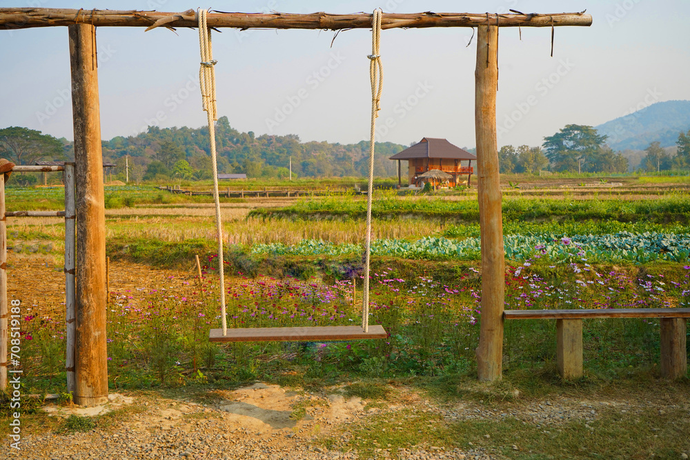 A wooden swing sits next to a flower garden and vegetable plot in a rice field.   