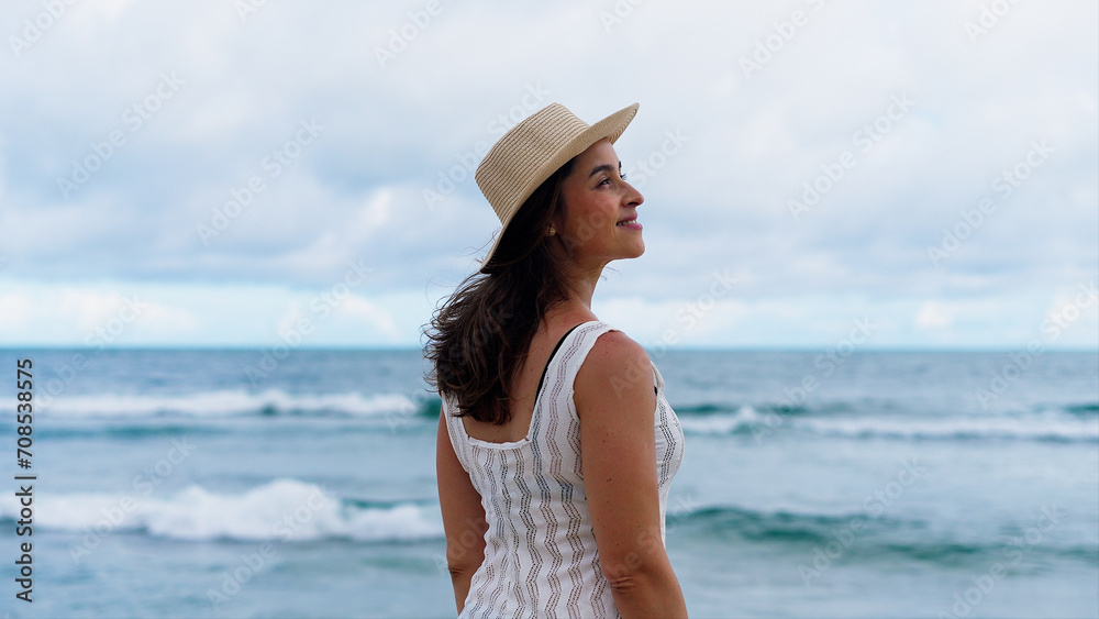 Happy young white woman wearing hat feeling the sea breeze. Beautiful smiling girl relaxing and enjoy the sun at seaside. 
