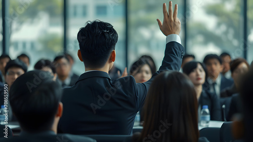 Asian businessman raises his hand to ask a question at a seminar speaker, wearing a suit, other listeners sit and listen, view from behind.
