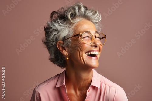 portrait of a beautiful elderly woman 60 years old in profile with a short haircut and glasses, laughs loudly, showing healthy teeth, in a pink blouse on a plain background in the studio