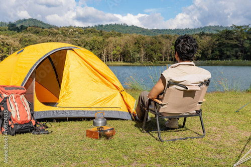 An Asian man sitting and drinking coffee near camping tent beside the lake and mountain in the sunshine morning with backpack and mobile solar cell