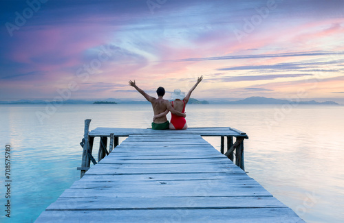 Romantic couple with arms up sitting on old wooden pier at lake, sunset shot