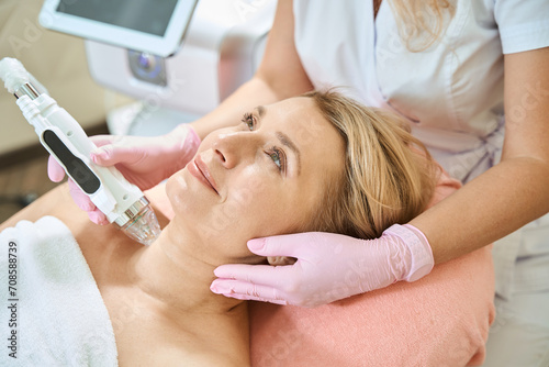 Cropped cosmetologist doing microneedle rf lifting of neck skin of smiling woman