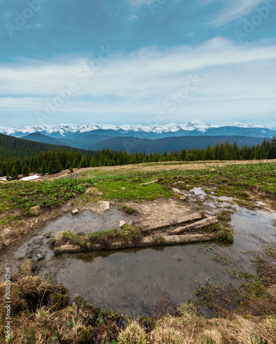 Spring Carpathian mountains landscape with snow-covered tops of Chornohora ridge in far, Ukraine. Fenced farming area with water spring and drinker.