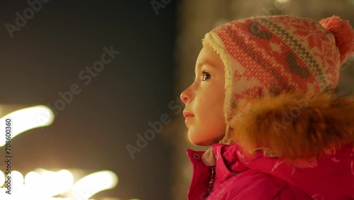 Portrait of a little girl in warm winter clothes outside on a cold Christmas night among beautiful lights, looking into the distance with dreamy eyes and making a wish. Steam is coming out of my mouth