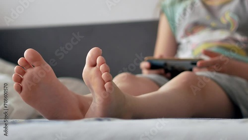 The bare feet of a little girl's child in close-up, sitting in bed with a smartphone in his hands.