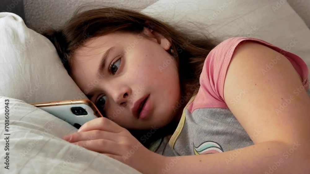 A little girl is sick in the morning, lying in bed in pajamas with disheveled hair watching videos on her smartphone. Weakness, a feeling of illness, sadness. A little girl is sick in the morning, lying in bed in pajamas with disheveled hair watching videos on her smartphone. Weakness, a feeling of illness, sadness.