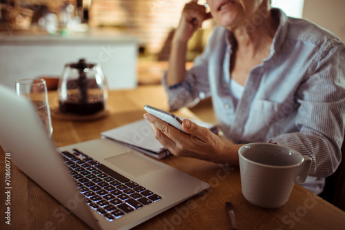 Senior woman using smartphone and laptop at home with coffee