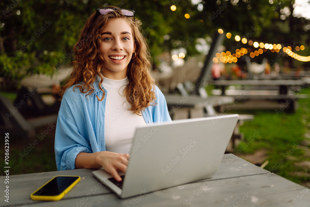 Young woman in stylish clothes sitting at a table outdoors working on a laptop with a beautiful view of the lake. Freelancing concept. Online education. Young woman in stylish clothes sitting at a table outdoors working on a laptop with a beautiful view of the lake. Freelancing concept. Online education.