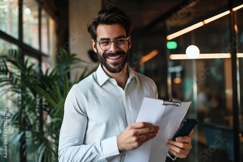 Smiling happy young bearded Latin professional business man executive holding documents and cell phone making mobile call at work on cellphone consulting client standing in modern, Generative AI