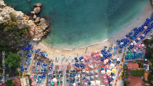 Top down view of sandy beach with turquoise sea water, colorful umbrellas and swimming people. Crashing waves in golden beach at Büyük Çakıl, Kaş resort in Turkey. Drone shot, Aerial view