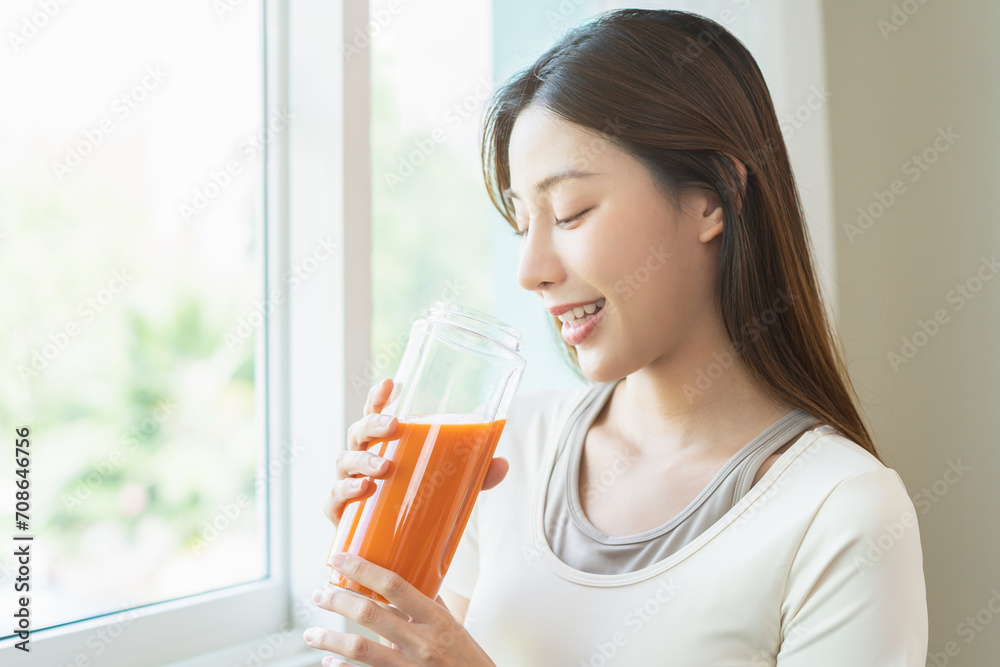 Orange detox juice, smile asian young woman hand holding glass of vegetable juice, carrot smoothie for diet near window at home, drinking healthy meal food for weight loss. Lifestyle, vegan nutrition. Orange detox juice, smile asian young woman hand holding glass of vegetable juice, carrot smoothie for diet near window at home, drinking healthy meal food for weight loss. Lifestyle, vegan nutrition.