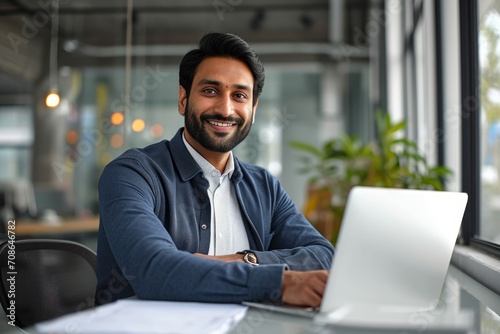 Smiling confident Indian business man employee looking at camera sitting at work desk with laptop computer. Portrait of smart happy businessman or entrepreneur posing at workplace, Generative AI © Design Dynasty