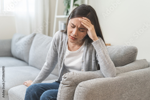 Dizzy asian young woman, girl headache or migraine pain suffering from vertigo while sitting on couch in living room at home, holding head with hand, health problem of brain or inner ear not balance. photo