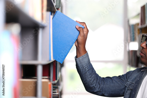 An African American man in a denim shirt picks out a book in a library or bookstore. Dark-skinned man in glasses choosing interesting book literature while standing in bookstore
