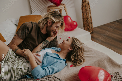 Loving couple embracing and smiling while lying in bed surrounded with red heart shape balloons