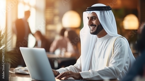 An Emirati man smiles while using his laptop in a modern workplace,