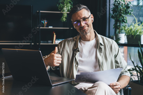 Portrait of Caucasian male employee in earbuds holding paper documents and smiling at camera in office interior, successful businessman in classic spectacles posing at desktop with laptop computer