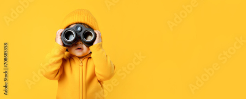 A cheerful baby looks through binoculars on a yellow background. Banner, copyspace