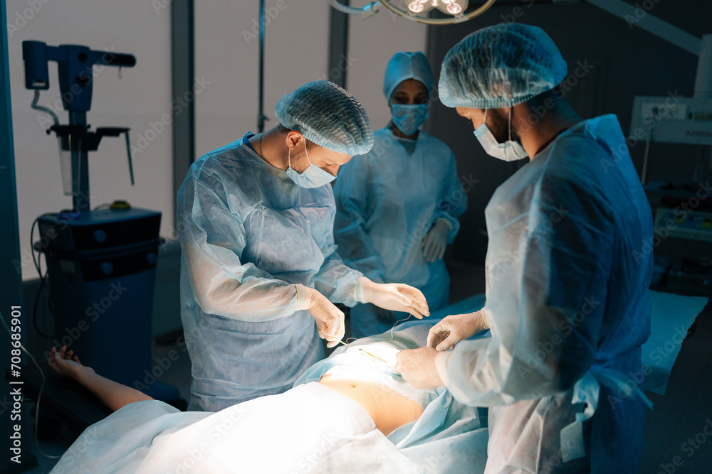 Remote view diverse team of professional surgeons performing invasive surgery on patient in hospital. African-American female assistant helping head surgeon apply sutures. Concept of emergency. Remote view diverse team of professional surgeons performing invasive surgery on patient in hospital. African-American female assistant helping head surgeon apply sutures. Concept of emergency.
