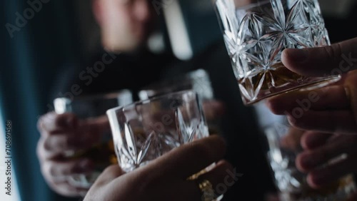hand holding a glass of whiskey. The person is wearing a white shirt with a cufflink visible. Another hand with a glass is in the background. The focus is on the glass in the foreground, suggesting  photo