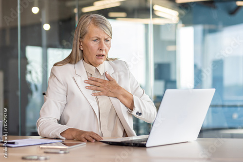 Senior woman in business suit sitting at desk in office in front of laptop and holding hand to chest, having panic attack, heart attack, shocked by business affairs.