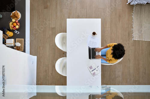 Young African American woman using laptop computer technology sitting at home kitchen table hybrid working online, female student elearning, searching on web typing at pc. Above overhead top view.