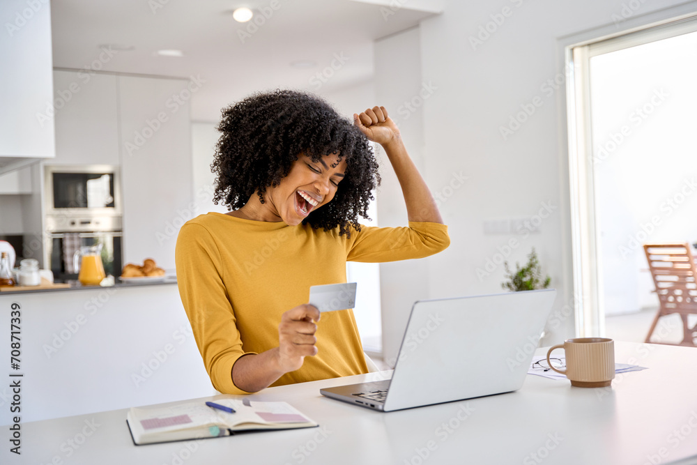 Happy excited young African woman customer winner holding credit card using laptop computer in kitchen celebrating money win receiving cashback getting online bonus for shopping sitting at home. - obrazy, fototapety, plakaty 