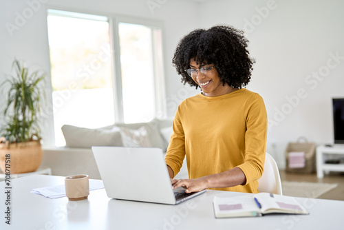 Young pretty happy smiling African American woman wearing eyeglasses using laptop computer sitting at home table hybrid working online, female student elearning, browsing web typing at pc. photo