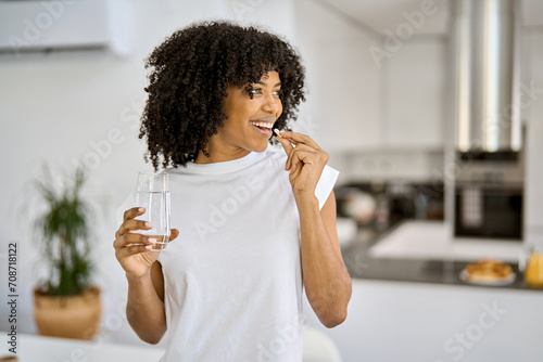 Happy healthy young adult African American woman model talking pill holding glass of water standing in the kitchen at home. Morning supplements vitamins nutrition treatment concept.