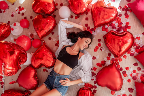 Top view of young woman lying on wooden floor surrounded by red heart shaped balloons in Valentines Day. Girl is smiling, wearing casual outfit. Floor with rose petals, romantic festive ambiance