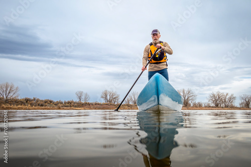 senior male paddler is paddling a stand up paddleboard on a calm lake in spring, frog perspective from an action camera at water level photo