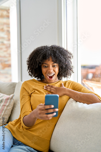 Excited young African American woman customer looking at mobile phone sitting on sofa at home. Happy lady using smartphone surprised about money win, lottery prize, shopping cashback or gift. Vertical