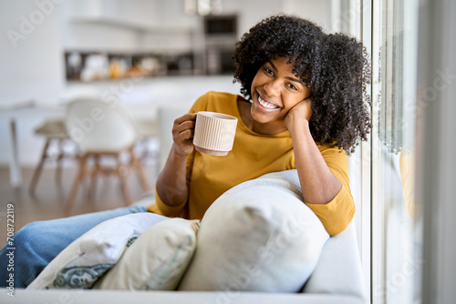 Happy young African woman sitting on comfortable sofa in living room at home. Smiling pretty girl drinking coffee relaxing on couch looking at camera in modern cozy house. Portrait.