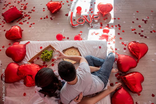 Top view of young couple sharing casual, intimate moment together on bed with red heart shaped balloons. Man and woman embracing each others in Valentines Day. Lovers enjoying pizza from takeaway box