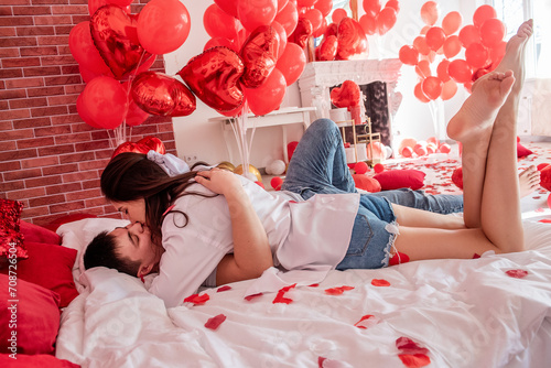 Couple in love hugs tightly on white bed among red balloons, pillows, hearts, rose petals. Man and woman look at each other, spend Valentines Day home in relaxed atmosphere. Surprise gift. Lifestyle photo