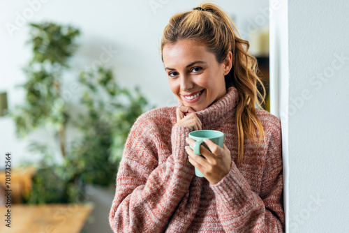 Beautiful young woman drinking a hot cup of coffee while looking at camera in living room at home.